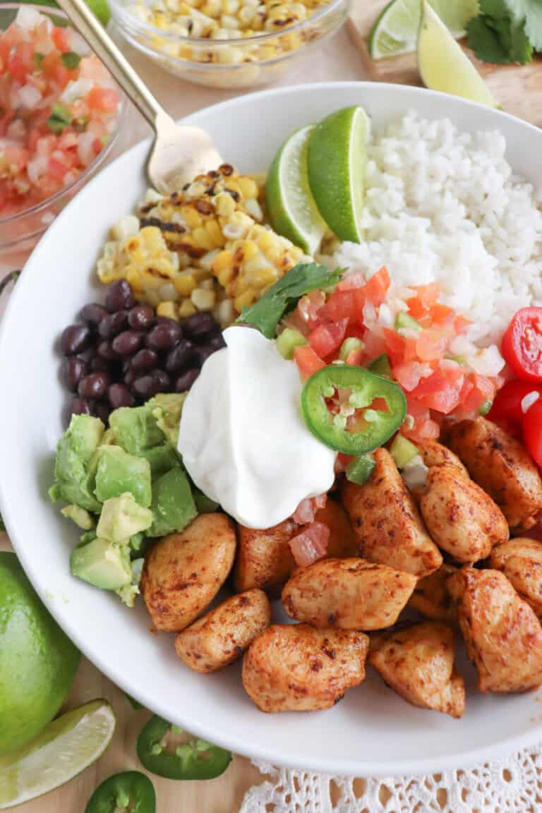 Cooked chicken bites with seasoning in a large white bowl with rice, diced avocado, black beans, corn kernels, cherry tomatoes, pico and sour cream topped with a slice of jalapeno and lime.