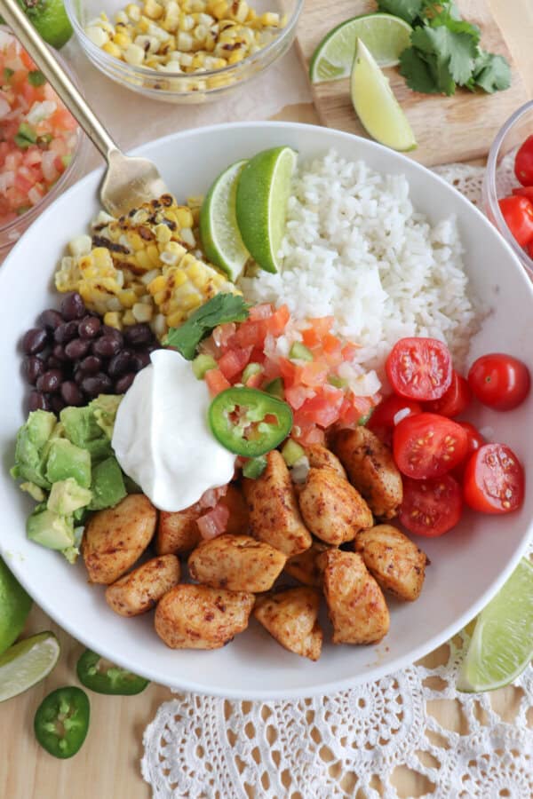 Cooked chicken bites with seasoning in a large white bowl with rice, diced avocado, black beans, corn kernels, cherry tomatoes, pico and sour cream topped with a slice of jalapeno and lime.