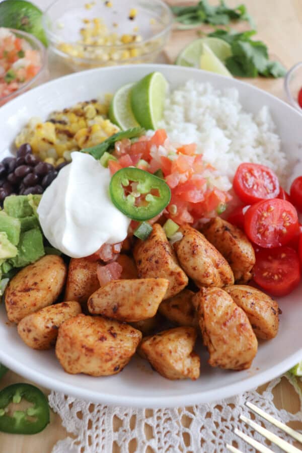 Cooked chicken bites with seasoning in a large white bowl with rice, diced avocado, black beans, corn kernels, cherry tomatoes, pico and sour cream topped with a slice of jalapeno and lime.