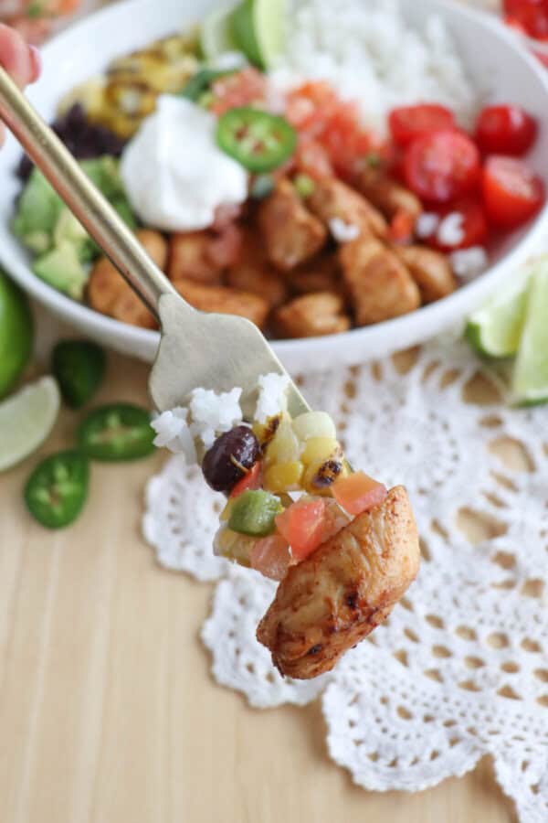 Cooked chicken bites with seasoning in a large white bowl with rice, diced avocado, black beans, corn kernels, cherry tomatoes, pico and sour cream topped with a slice of jalapeno and lime.