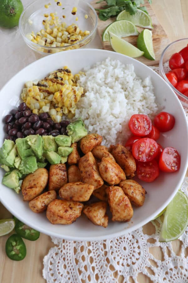 Cooked chicken bites with seasoning in a large white bowl with rice, diced avocado, black beans, corn kernels and cherry tomatoes.
