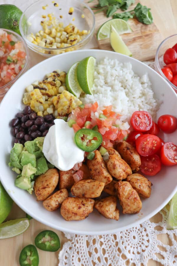 Cooked chicken bites with seasoning in a large white bowl with rice, diced avocado, black beans, corn kernels, cherry tomatoes, pico and sour cream topped with a slice of jalapeno and lime.