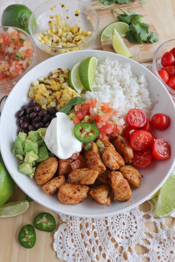 Cooked chicken bites with seasoning in a large white bowl with rice, diced avocado, black beans, corn kernels, cherry tomatoes, pico and sour cream topped with a slice of jalapeno and lime.