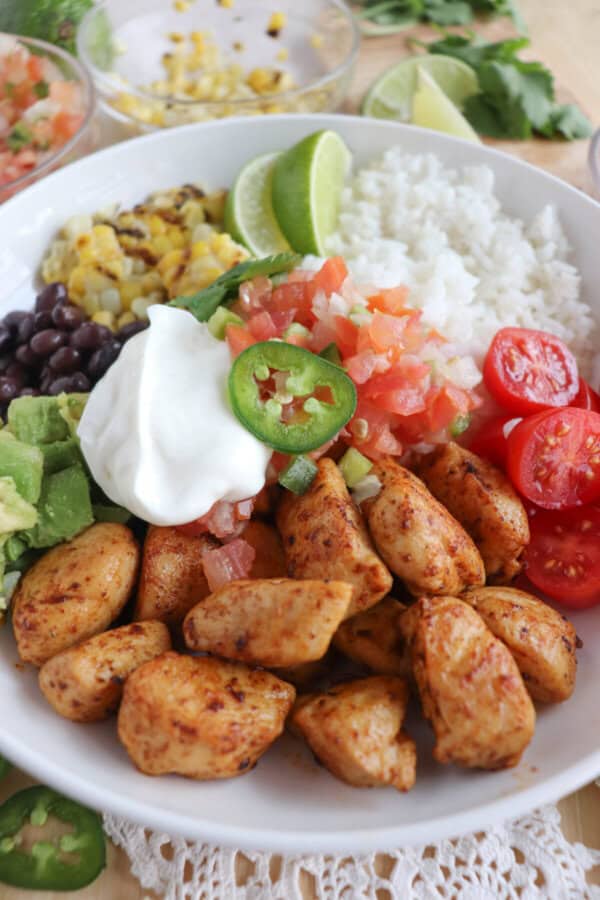 Cooked chicken bites with seasoning in a large white bowl with rice, diced avocado, black beans, corn kernels, cherry tomatoes, pico and sour cream topped with a slice of jalapeno and lime.