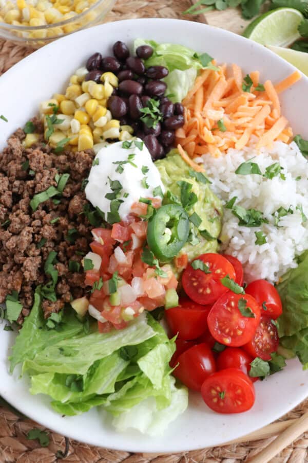 Large white bowl filled with cooked ground taco beef, rice, pico, corn, beans, cheddar cheese and cherry totmatoes, topped with guacamole and sour cream.
