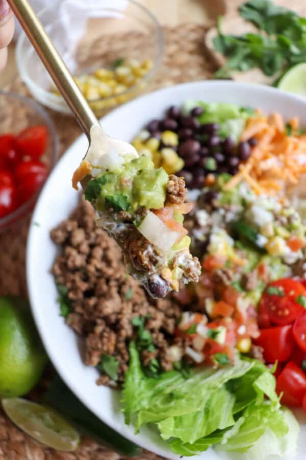 Large white bowl filled with cooked ground taco beef, rice, pico, corn, beans, cheddar cheese and cherry totmatoes, topped with guacamole and sour cream with a fork taking a bite.