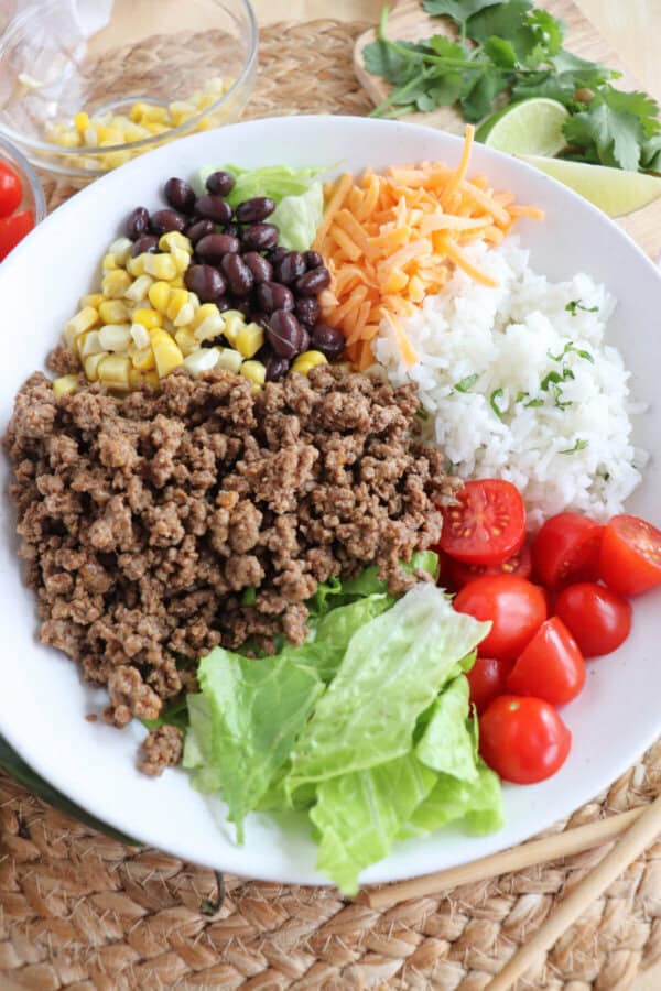 Ground beef, rice, corn, beans, shredded cheese, halved cherry tomatoes and lettuce in a large white bowl.