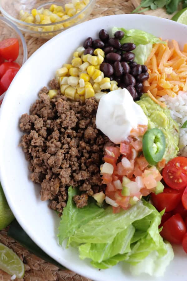 Large white bowl filled with cooked ground taco beef, rice, pico, corn, beans, cheddar cheese and cherry totmatoes, topped with guacamole and sour cream.