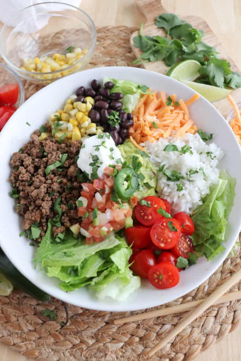 Large white bowl filled with cooked ground taco beef, rice, pico, corn, beans, cheddar cheese and cherry totmatoes, topped with guacamole and sour cream.