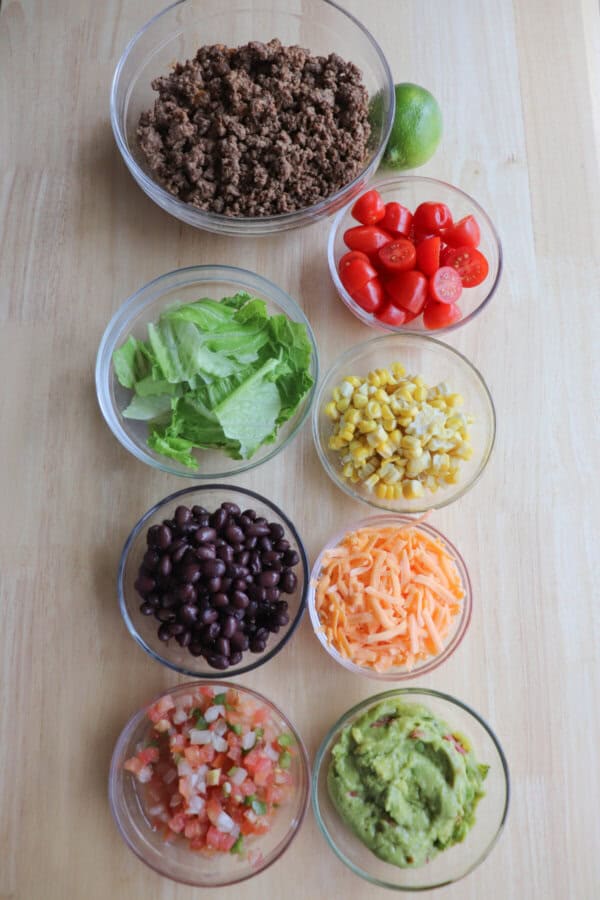 Ingredients for ground beef burrito bowls in clear glass bowls on a wooden table.