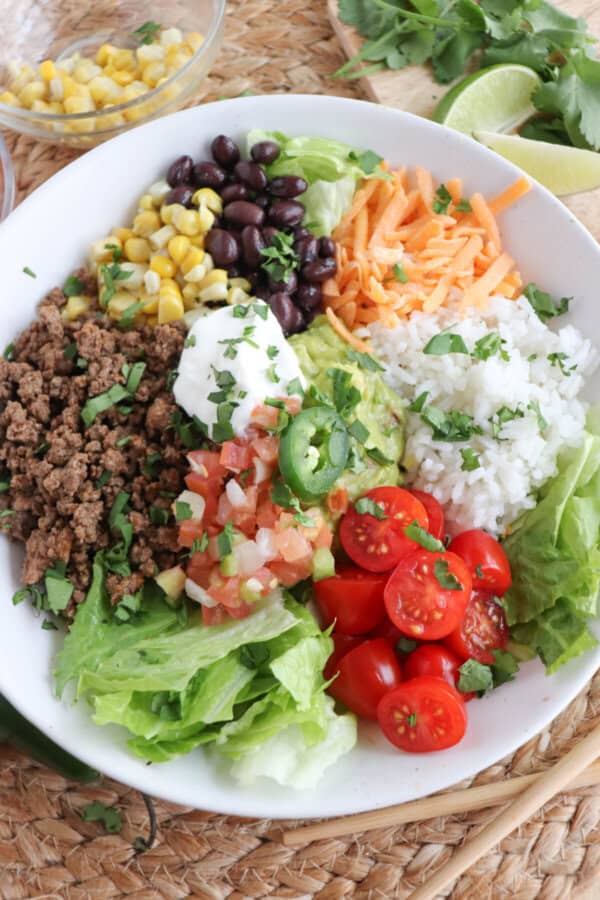 Large white bowl filled with cooked ground taco beef, rice, pico, corn, beans, cheddar cheese and cherry totmatoes, topped with guacamole and sour cream.