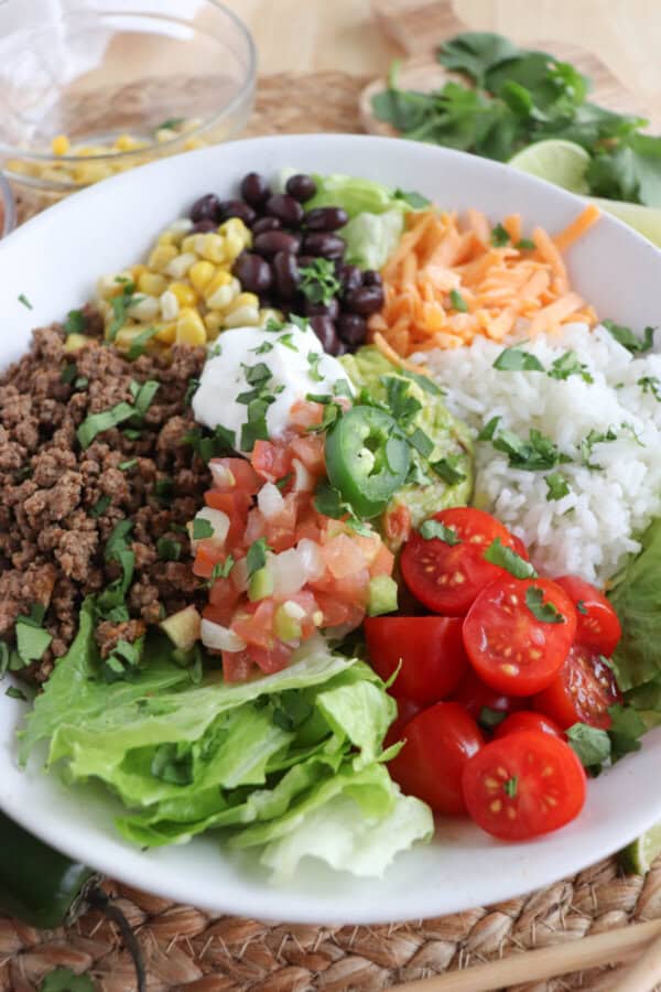 Large white bowl filled with cooked ground taco beef, rice, pico, corn, beans, cheddar cheese and cherry totmatoes, topped with guacamole and sour cream.