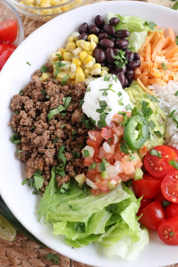 Large white bowl filled with cooked ground taco beef, rice, pico, corn, beans, cheddar cheese and cherry totmatoes, topped with guacamole and sour cream.