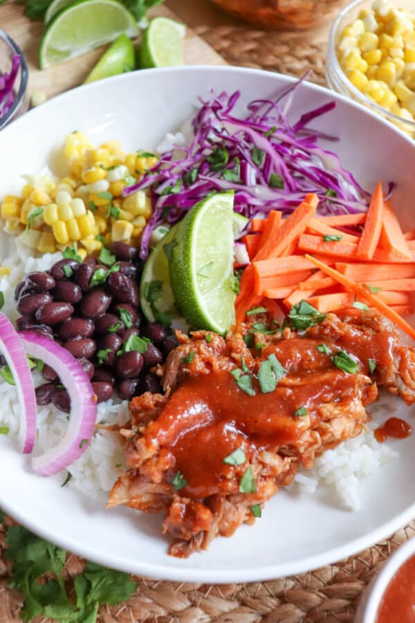 Pulled pork and vegetables over rice in a large white bowl topped with barbecue sauce.