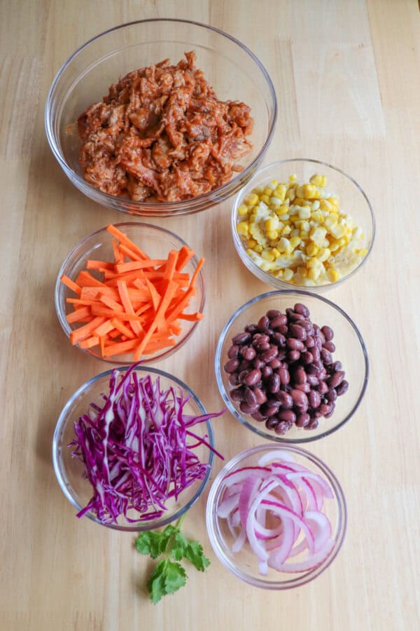 Pulled pork burrito bowl ingredients in clear glass bowls on a wooden table.