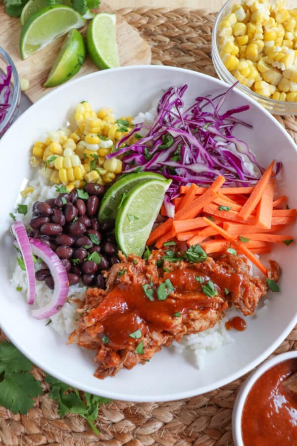 Pulled pork and vegetables over rice in a large white bowl topped with barbecue sauce.