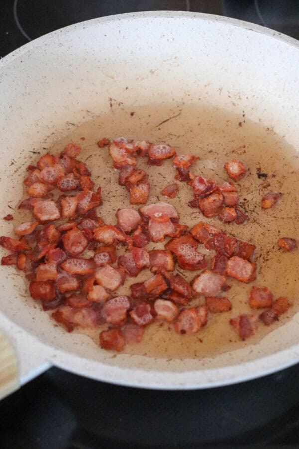 Bacon bits cooking on a stove top in a white skillet.