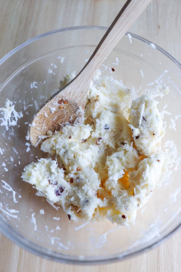 Butter, cheese and herbs in a clear glass bowl with a wooden spoon.