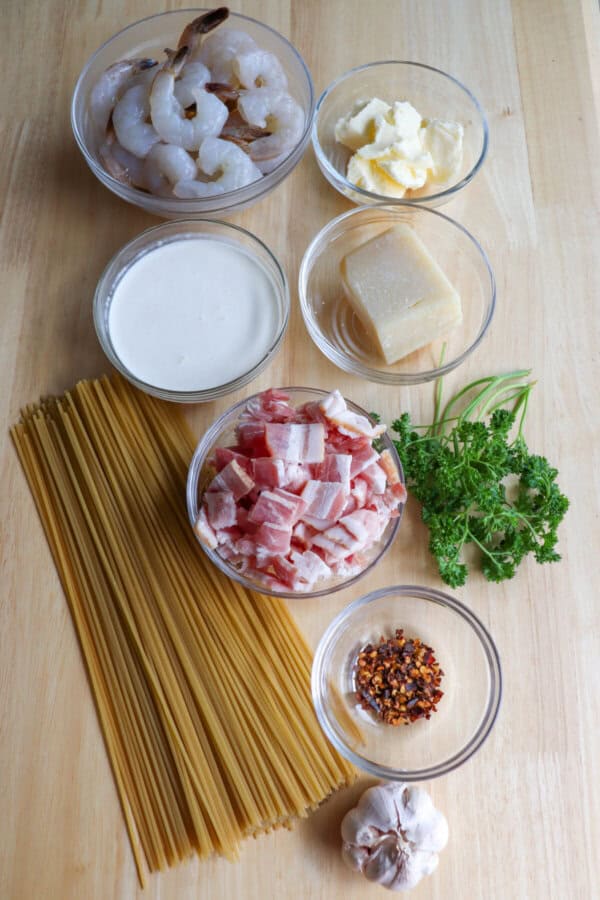 Ingredients to make shrimp spaghetti in clear glass bowls on a wooden table.