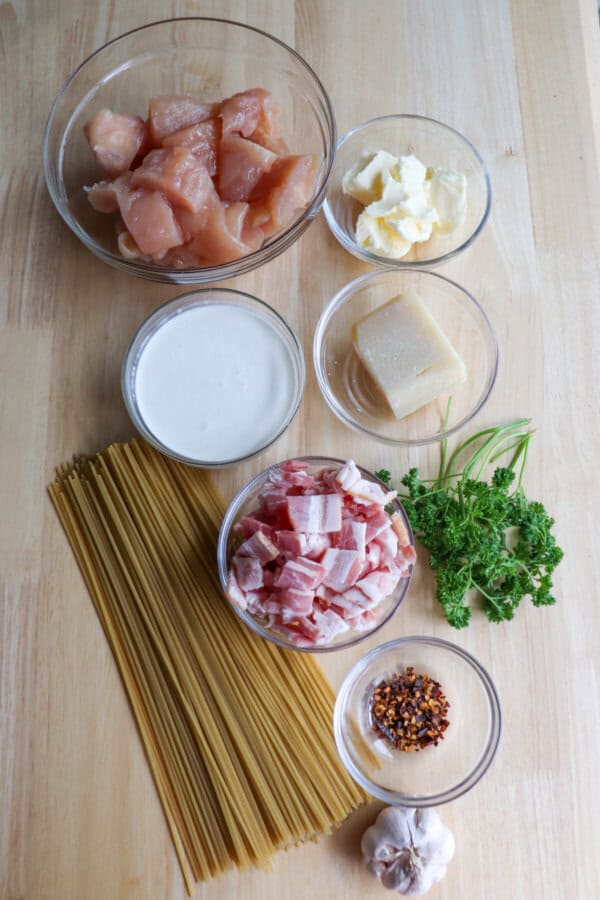 Ingredients for chicken spaghetti in clear glass bowls on a wooden table.