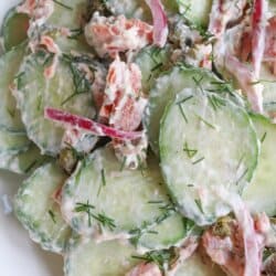 Sliced cucumbers with pieces of smoked salmon and pickled onions covered in a creamy dill sauce in a white bowl.