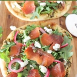 Small round pizza on a wooden cutting board topped with fresh arugula, smoked salmon, goat cheese and pickled onions.
