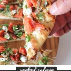 A flatbread topped with smoked salmon, mustard greens, goat cheese and balsamic vinegar, cut into strips on a wooden board with one being held in a hand.