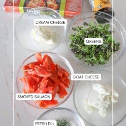 Ingredients for smoked salmon flatbread in clear glass bowls on a white counter.