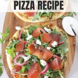 Small round pizza on a wooden cutting board topped with fresh arugula, smoked salmon, goat cheese and pickled onions.