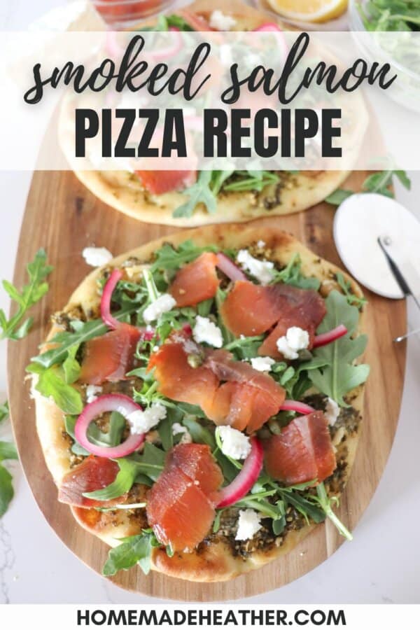 Small round pizza on a wooden cutting board topped with fresh arugula, smoked salmon, goat cheese and pickled onions.