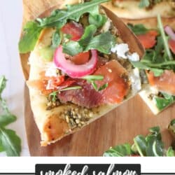 Piece of pizza above a wooden cutting board topped with fresh arugula, smoked salmon, goat cheese and pickled onions.