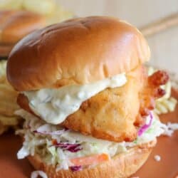 Fried fish filet on a brioche bun with coleslaw and tartar sauce on an orange plate.