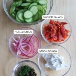 Cucumber smoked salmon salad ingredients in clear glass bowls on a wooden surface.