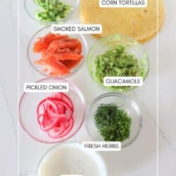 Ingredients for smoked salmon tacos in clear glass bowls on a white counter.