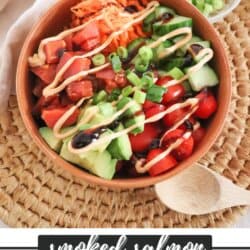 An orange bowl filled with smoked salmon, shredded carrots, cucumber slices, cherry tomatoes, avocado, green onions, and drizzled with sauces, creating a visually appetizing and healthy dish.