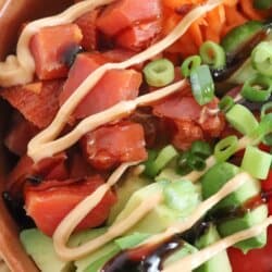 An orange bowl filled with smoked salmon, shredded carrots, cucumber slices, cherry tomatoes, avocado, green onions, and drizzled with sauces, creating a visually appetizing and healthy dish.
