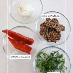 Ingredients for smoked salmon crackers in clear glass bowls on a white surface.