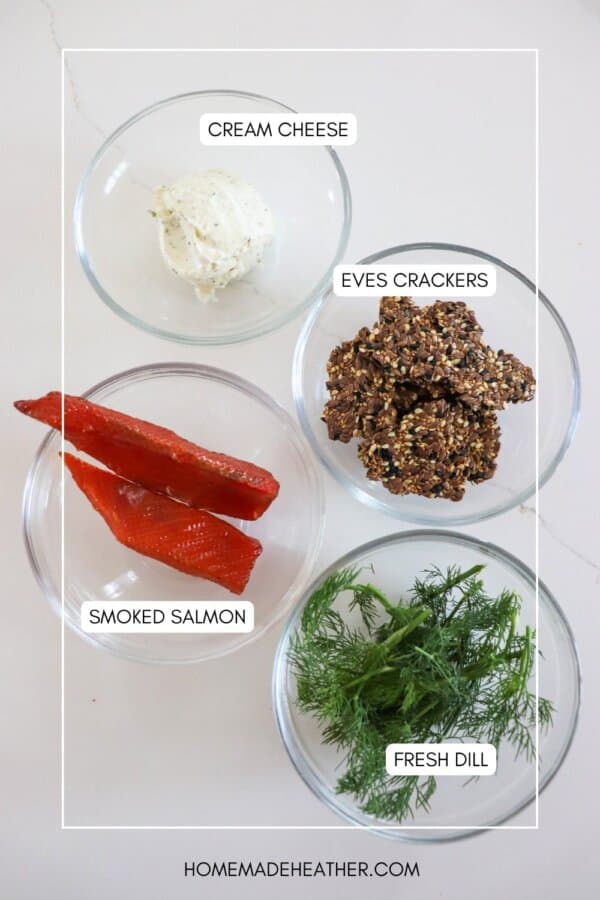 Ingredients for smoked salmon crackers in clear glass bowls on a white surface.