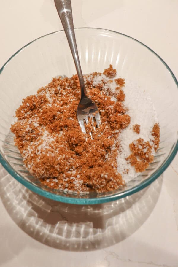 Coarse salt and sugar being mixed in a clear glass bowl with a fork.