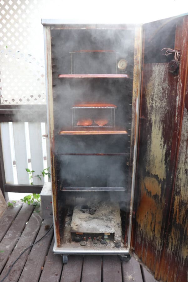 Smoke billowing out from a vertical smoker with salmon fillets inside.