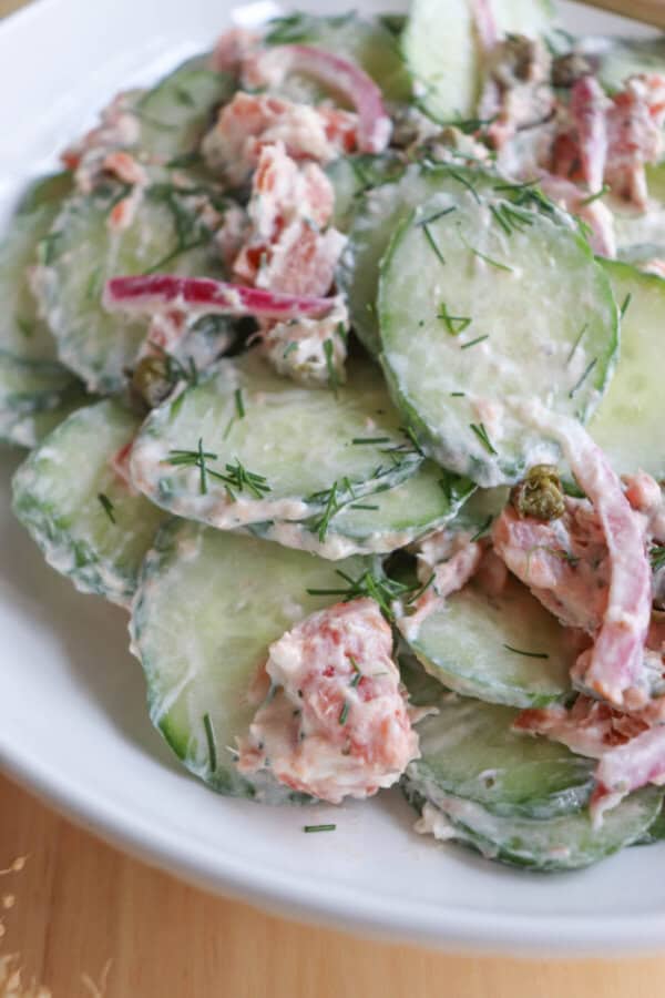 Sliced cucumbers with pieces of smoked salmon and pickled onions covered in a creamy dill sauce in a white bowl.
