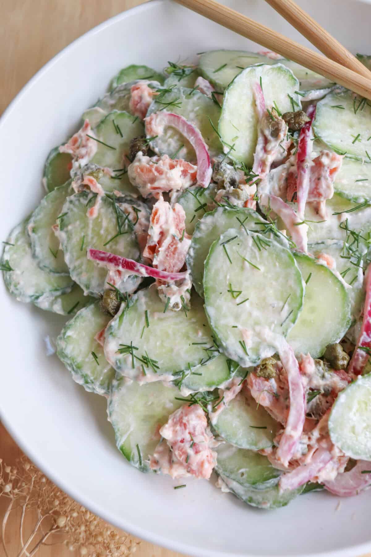 Sliced cucumbers with pieces of smoked salmon and pickled onions covered in a creamy dill sauce in a white bowl.