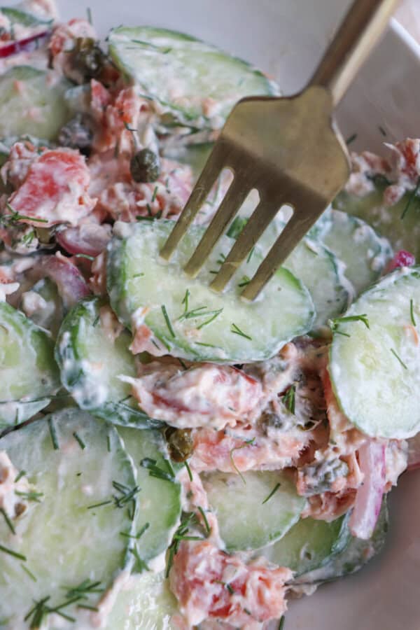Sliced cucumbers with pieces of smoked salmon and pickled onions covered in a creamy dill sauce with a gold fork.