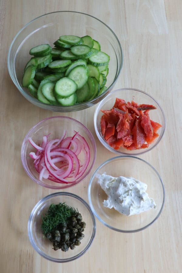 Cucumber smoked salmon salad ingredients in clear glass bowls on a wooden surface.