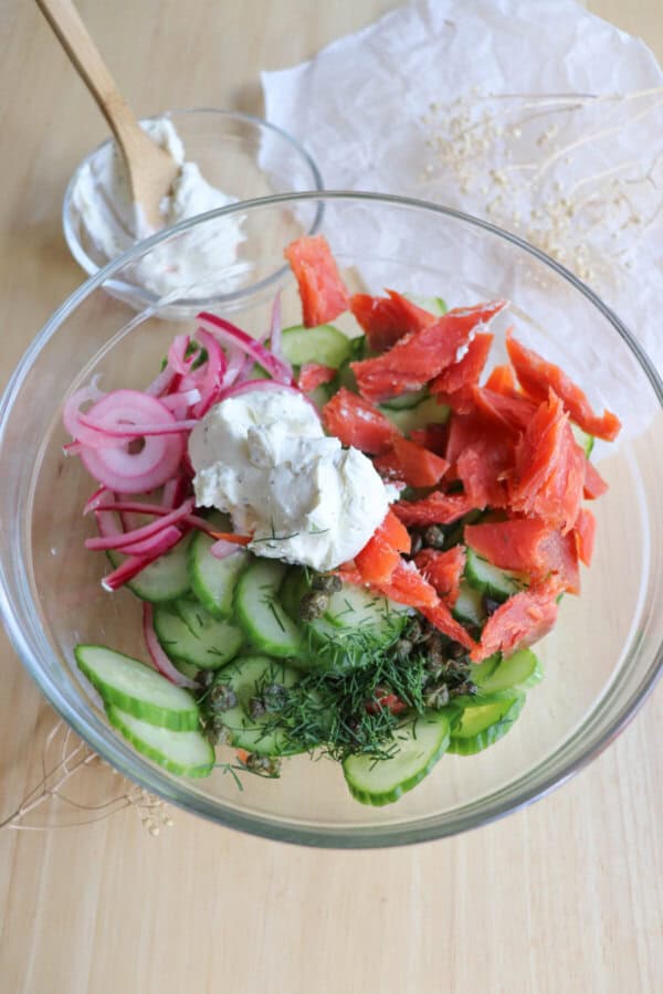 Sliced cucumber, smoked salmon pieces, fresh dill and pickled onion pieces in a clear glass mixing bowl.