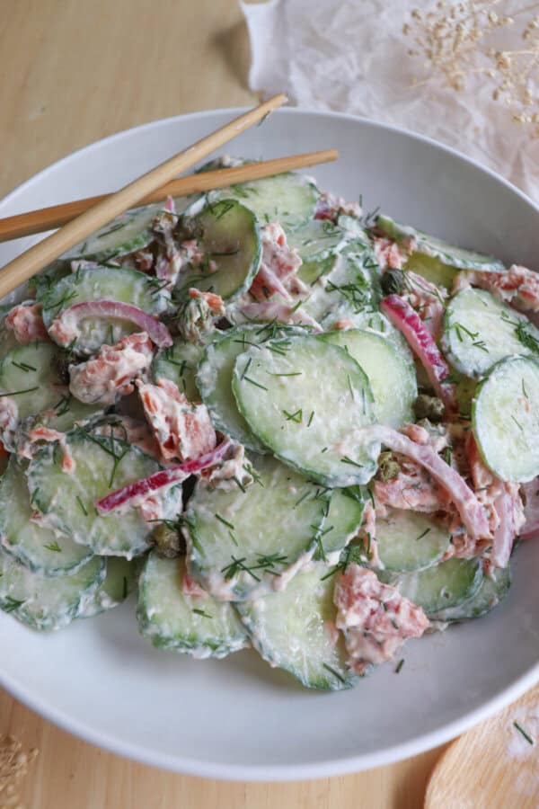 Sliced cucumbers with pieces of smoked salmon and pickled onions covered in a creamy dill sauce in a white bowl.