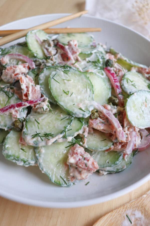 Sliced cucumbers with pieces of smoked salmon and pickled onions covered in a creamy dill sauce in a white bowl.