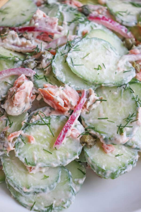 Sliced cucumbers with pieces of smoked salmon and pickled onions covered in a creamy dill sauce in a white bowl.