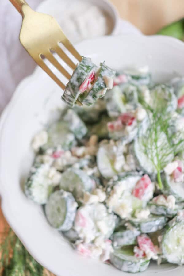 Sliced cucumbers in tzatziki with diced pickled onions and dill in a white bowl with a gold fork with cucumbers on it.