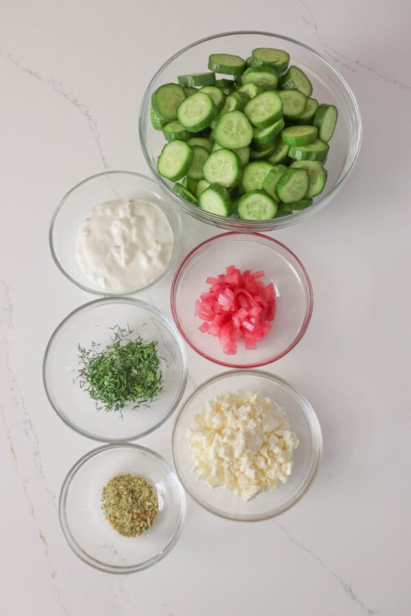 Ingredients for tzatziki cucumber salad in clear glass bowls on a white counter.